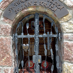 Kovaný pamätník svätých s atribútmi na mrežiach. Sv. Terézia Avilská - srdce s nápisom IHS, Sv. Ján z Kríža - kríž